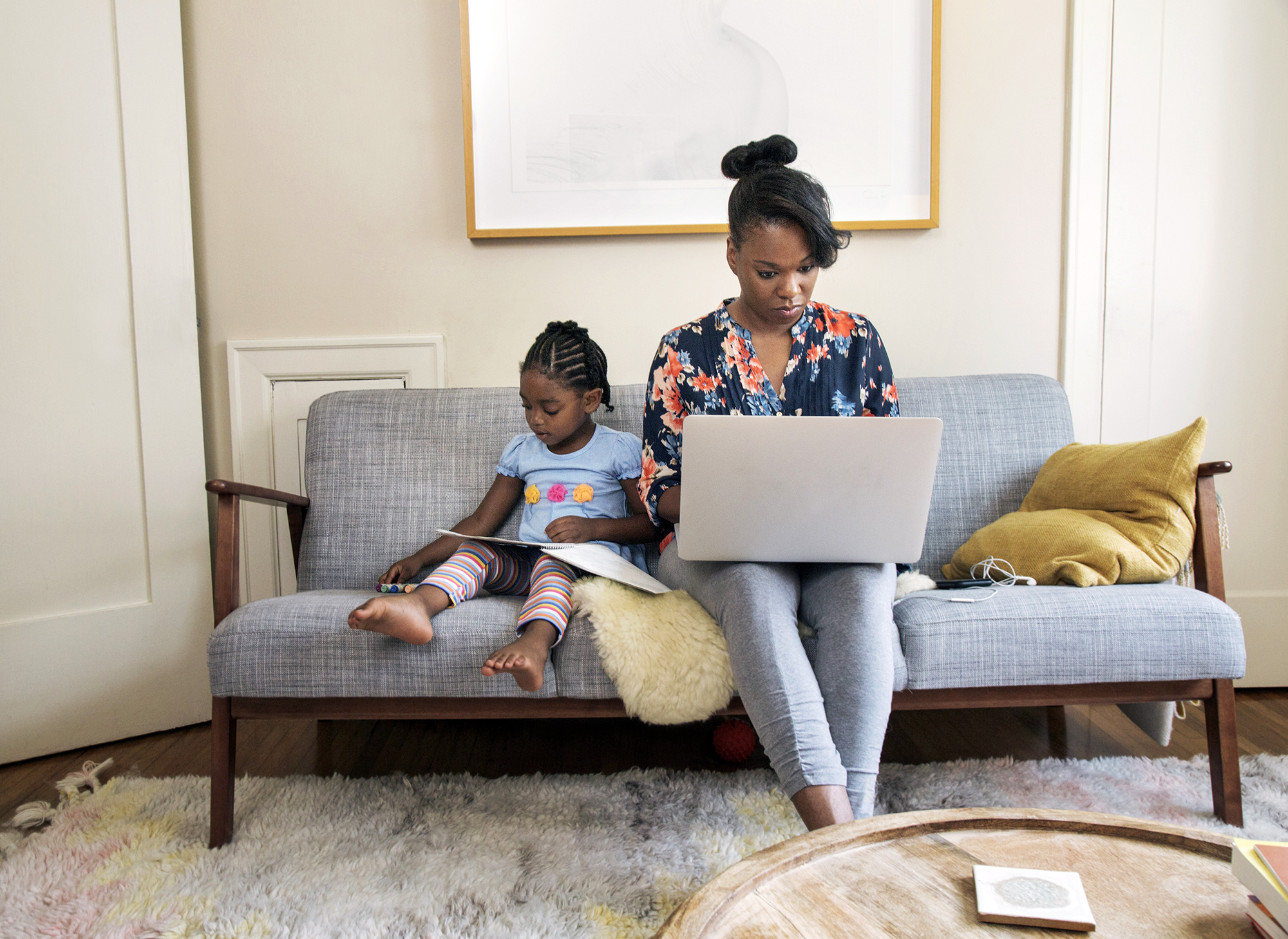 A Black woman sits on a gray couch working on a laptop while her young child beside her uses a tablet. They are both focused on their devices in a cozy, well-lit living room with pillows and a rug.