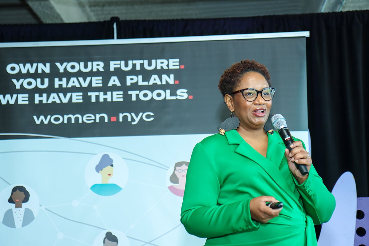 A person in a green blazer speaks into a microphone at an event with a sign reading "Own your future. You have a plan. We have the tools. women.nyc."