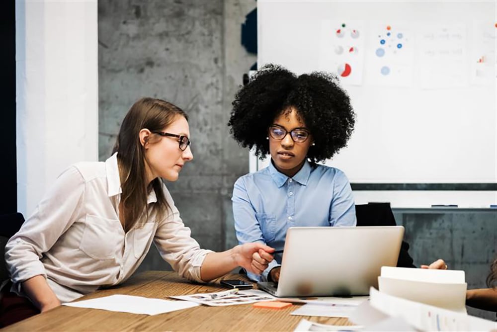 Two women sit at a desk working together, looking at a laptop. One woman gestures toward the screen while the other listens. Papers and a notebook are on the table; a whiteboard with charts is visible in the background.