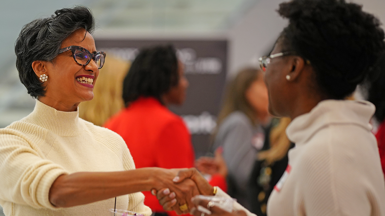 Two women smiling and shaking hands at an indoor event. One wears glasses and a white turtleneck, the other wears a cream sweater, with a blurred group of people in the background.