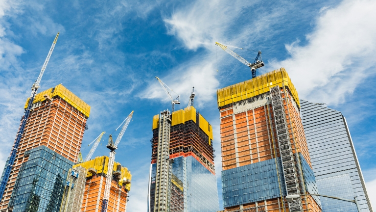 Tall skyscrapers under construction in Hudson Yards with cranes on top against a blue sky with white clouds. The buildings have glass and steel exteriors, with yellow and orange safety netting around upper floors.