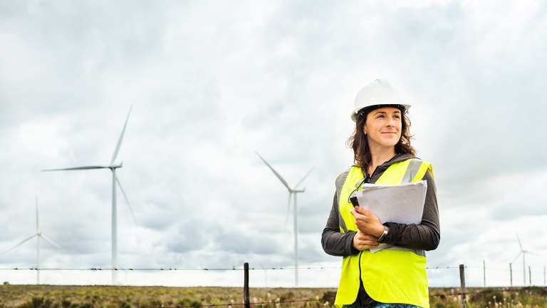 Smiling young female engineer in protective workwear standing by turbines during an inspection at a wind farm.