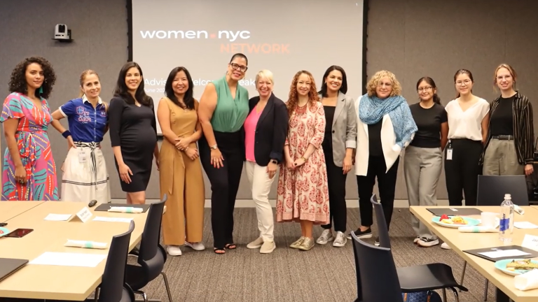 A group of eleven people stands in front of a presentation screen displaying &quot;women.nyc NETWORK.&quot; They are smiling and dressed in casual to business-casual attire.