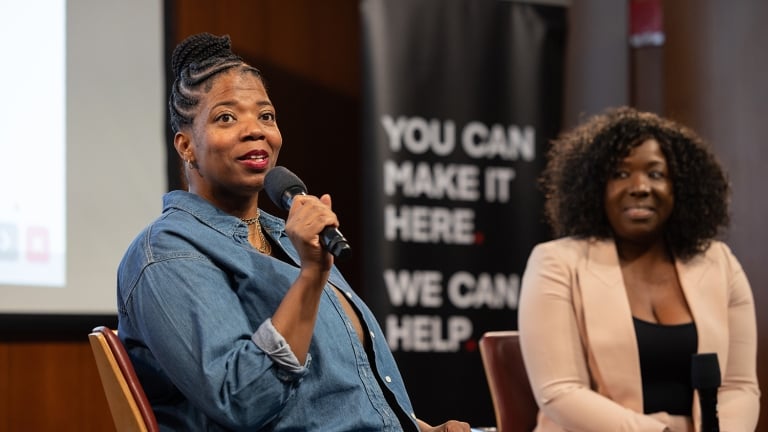 Two women are seated and holding microphones during a speaking event. One wears a denim top, while the other is dressed in a light blazer. A banner in the background reads, &quot;You can make it here. We can help.&quot; 