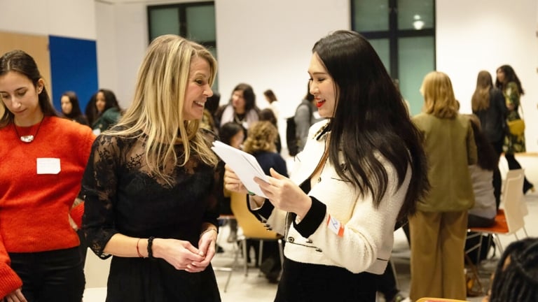 Two women are engaged in a conversation, one holding a piece of paper. They are in a well-lit room with other people and chairs in the background.