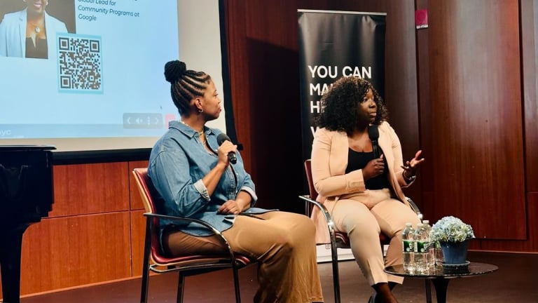 Two women seated on stage, engaged in discussion, with a large screen displaying visuals behind them.