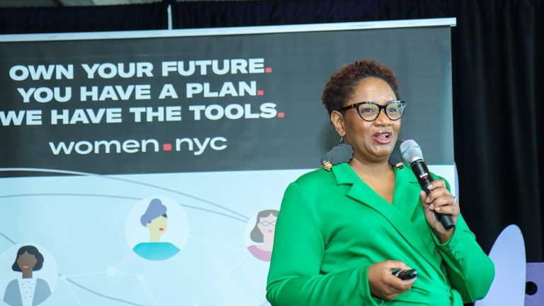 A person in a green blazer speaks into a microphone at an event with a sign reading "Own your future. You have a plan. We have the tools. women.nyc." A person in a green blazer speaks into a microphone at an event with a sign reading "Own your future. You have a plan. We have the tools. women.nyc."