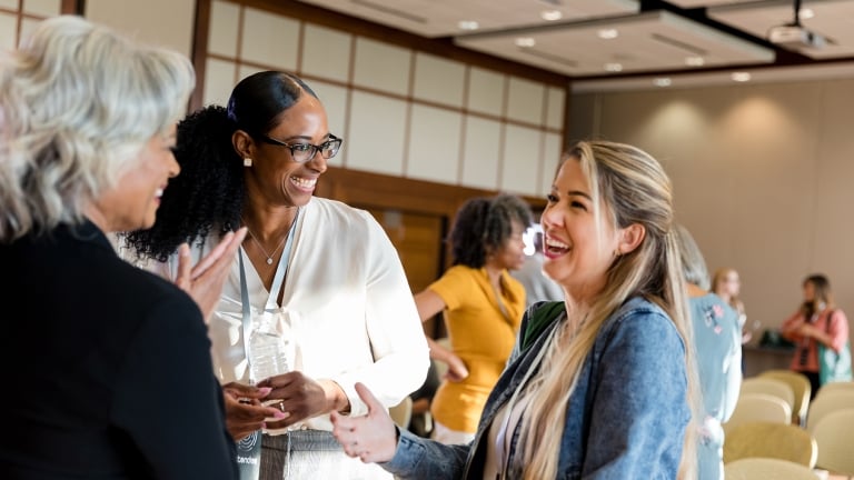 Three women are smiling and talking in a conference room with others in the background. They appear to be engaged in a friendly conversation, wearing casual business attire, and have conference badges. The room has a bright, professional atmosphere.