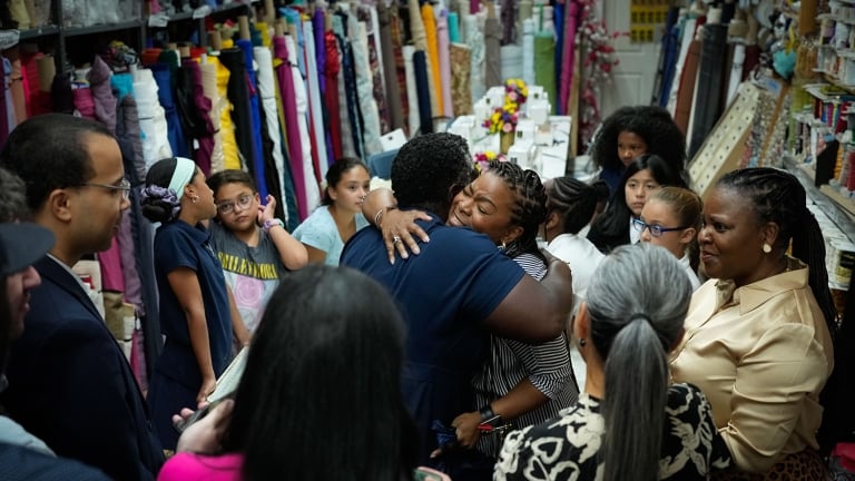 People are gathered in a fabric store, surrounded by colorful rolls of fabric. Two individuals are hugging warmly in the center, while others watch and smile. Shelves are filled with textile goods, creating a vibrant and lively atmosphere.