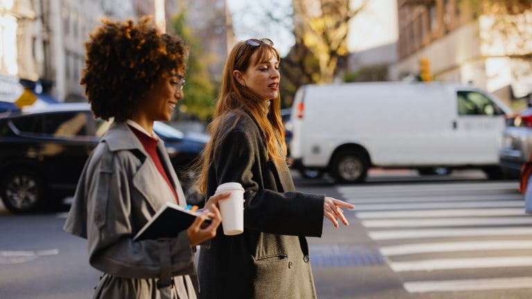 Two women walk across a city street crosswalk. One holds a coffee cup and notebook, while the other gestures as she speaks. Cars and buildings line the background on a sunny day.