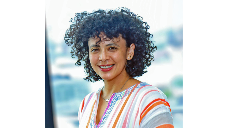 Sofia Mussa smiles at the camera with curly dark hair. She is wearing a striped, colorful top with white, orange, pink, and blue lines. The background is bright and blurry, suggesting a window with natural light.