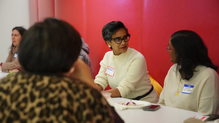 Three women sit around a table at a meeting, with two women wearing name tags facing each other while talking.