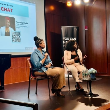 Two women seated on stage, engaged in discussion, with a large screen displaying visuals behind them.