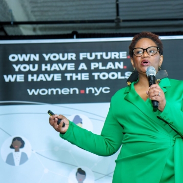 A person in a green blazer speaks into a microphone at an event with a sign reading &quot;Own your future. You have a plan. We have the tools. women.nyc.&quot;