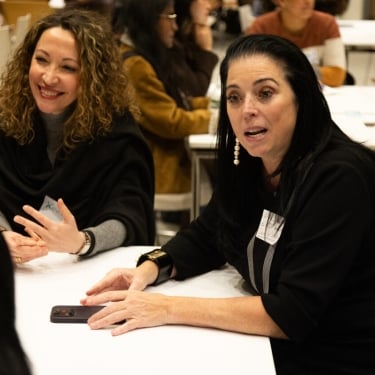 People are sitting and talking around a table in a social or meeting setting. Two women are in focus, engaged in conversation.