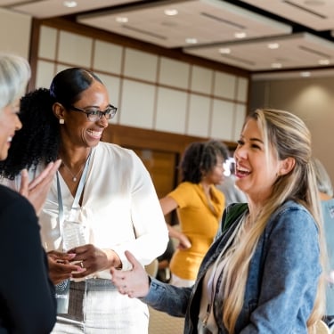 Three women are smiling and talking in a conference room with others in the background. They appear to be engaged in a friendly conversation, wearing casual business attire, and have conference badges. The room has a bright, professional atmosphere.