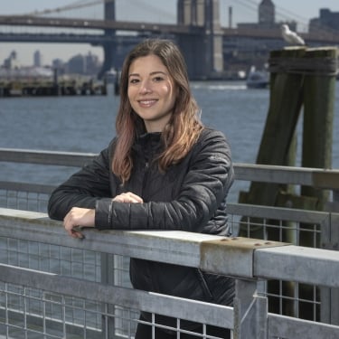 Bianca Mesqueue Reyes with long hair, wearing a black jacket, smiles while leaning on a metal railing. Behind her, the Brooklyn Bridge and New York City scape are visible across the water. It’s a clear day with a blue sky.