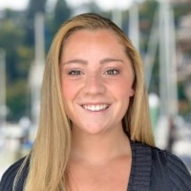 Smiling Brynna O’Leary with long blonde hair wearing a dark top, standing in front of a blurred marina backdrop with sailboats. Smiling Brynna O’Leary with long blonde hair wearing a dark top, standing in front of a blurred marina backdrop with sailboats.