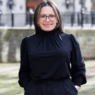 A smiling Nury M. Gutierrez in glasses stands outdoors in front of a historic building. She is wearing a black blouse and pants, with her hands in her pockets. The background features stone architecture and a grassy area. A smiling Nury M. Gutierrez in glasses stands outdoors in front of a historic building. She is wearing a black blouse and pants, with her hands in her pockets. The background features stone architecture and a grassy area.