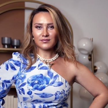 Irina Kanaeva with blonde hair wears a blue and white floral one-shoulder dress and pearl jewelry, standing confidently with hands on hips in a modern room with shelves and round glass lights in the background. Irina Kanaeva with blonde hair wears a blue and white floral one-shoulder dress and pearl jewelry, standing confidently with hands on hips in a modern room with shelves and round glass lights in the background.