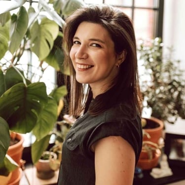 Joanna Kaniewska with straight brown hair, smiles while standing indoors next to large leafy plants and potted greenery, with natural light coming through a window in the background. Joanna Kaniewska with straight brown hair, smiles while standing indoors next to large leafy plants and potted greenery, with natural light coming through a window in the background.