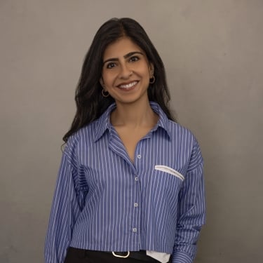 Shefali Lakhani with long dark hair smiles at the camera. She is wearing a blue and white striped button-up shirt and dark pants, standing against a plain, light gray background. Shefali Lakhani with long dark hair smiles at the camera. She is wearing a blue and white striped button-up shirt and dark pants, standing against a plain, light gray background.