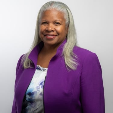 Deneen Donnley with long gray hair, wearing a purple blazer over a patterned top, smiling against a plain white background. Deneen Donnley with long gray hair, wearing a purple blazer over a patterned top, smiling against a plain white background.