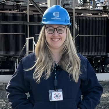 Kayla Mallay smiling outdoors and wearing glasses, a blue Con Edison hard hat, and a navy jacket. She has long blonde hair and a lanyard with an ID badge. Industrial equipment and structures are in the background. Kayla Mallay smiling outdoors and wearing glasses, a blue Con Edison hard hat, and a navy jacket. She has long blonde hair and a lanyard with an ID badge. Industrial equipment and structures are in the background.