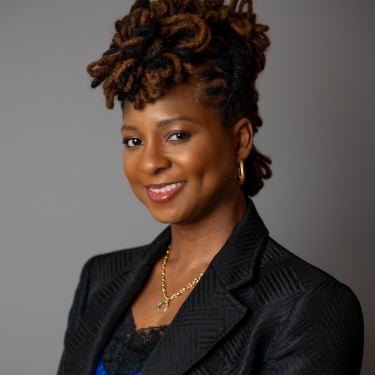 Nicole Mosby with styled locs, wearing a black blazer over a blue top, gold necklace, and hoop earrings, smiles at the camera against a plain gray background. Nicole Mosby with styled locs, wearing a black blazer over a blue top, gold necklace, and hoop earrings, smiles at the camera against a plain gray background.