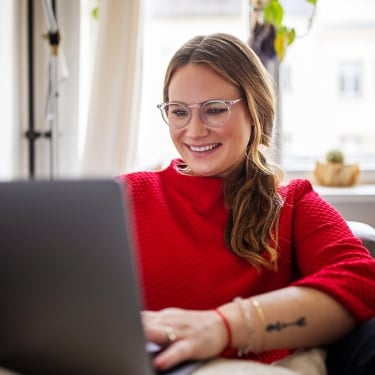 A woman wearing glasses and a red sweater is smiling while using a laptop, sitting comfortably indoors by a window with soft natural light.