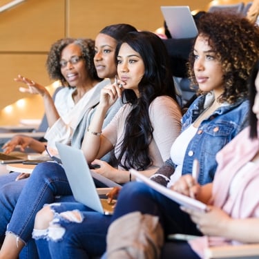 While her friends look at the unseen teacher in the front of the lecture hall, the mature adult female college student gestures as she speaks.he speaker and others writing.