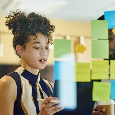 Two people stand by a glass wall covered with colorful sticky notes, discussing ideas. The woman in front holds a tablet and stylus, while the man behind her holds a coffee cup. The scene suggests teamwork or brainstorming.