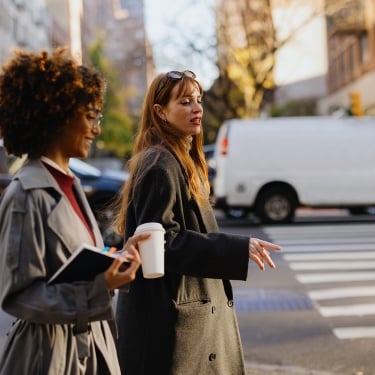 Two women walk across a city street crosswalk. One holds a coffee cup and notebook, while the other gestures as she speaks. Cars and buildings line the background on a sunny day.