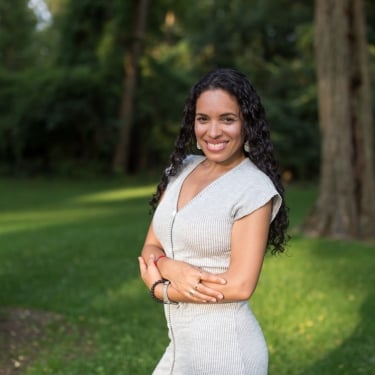 Woman with long curly hair, Aneika Perez, wearing a light gray dress, stands smiling with arms crossed in a sunlit park with green grass and tall trees in the background.