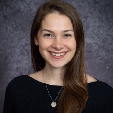 Kendall Thompson with long brown hair, wearing a black top and a round pendant necklace, smiles in front of a gray mottled background.