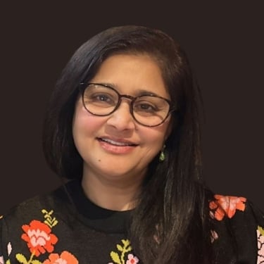 Apexa Dave with long dark hair and glasses smiles. She is wearing a black sweater with colorful floral embroidery on the shoulders. The background is solid dark brown.