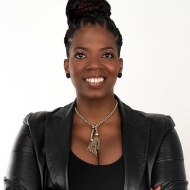 Regina Gwynn with dark braided hair in a bun, wearing a black leather jacket, black top, gold necklace, and stud earrings, stands with arms crossed and smiles confidently against a plain white background.