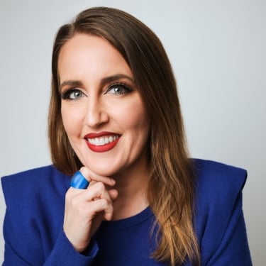 A woman with long brown hair and blue eyes, Stephanie Farquhar, wearing a bright blue top and matching ring, smiles while resting her chin on her hand against a light background.