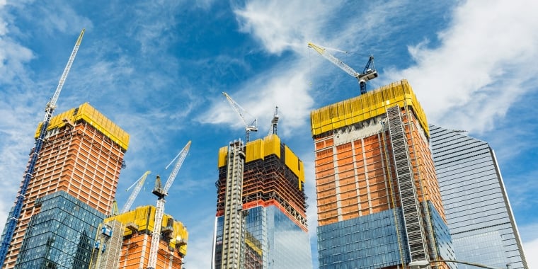 Tall skyscrapers under construction in Hudson Yards with cranes on top against a blue sky with white clouds. The buildings have glass and steel exteriors, with yellow and orange safety netting around upper floors.