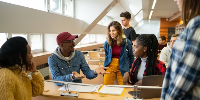 A group of students in a classroom engage in discussion, sitting and standing around desks with papers and a laptop.