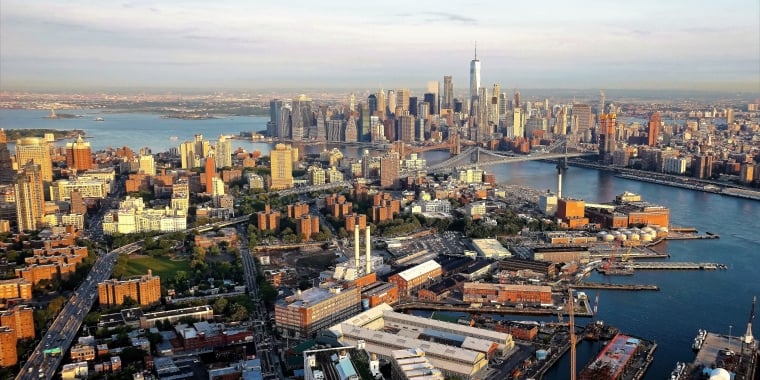 Aerial view of Manhattan, New York City, showcasing skyscrapers, the Hudson River, and surrounding boroughs. The One World Trade Center is prominently visible, with sunlit buildings and bustling waterways in the foreground.