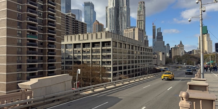 Urban scene at 100 Gold Street with a yellow taxi driving on a bridge in Manhattan. Tall skyscrapers are in the background beneath a partly cloudy sky. The setting appears busy and modern with multiple lanes on the road.