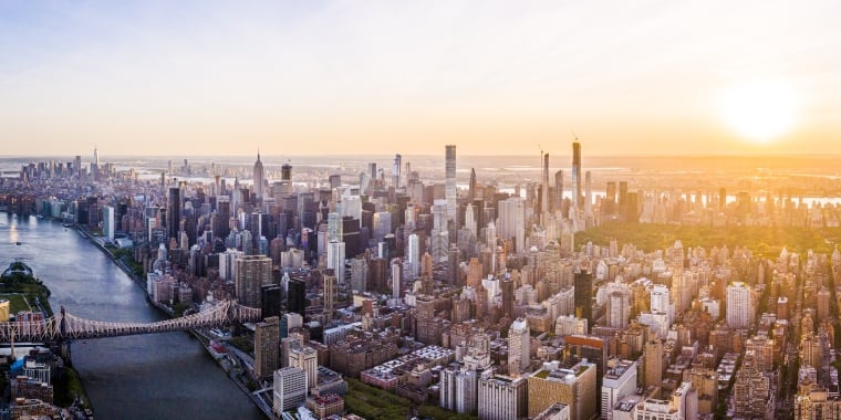 Aerial view of New York City skyline during sunset, featuring numerous skyscrapers and buildings. A wide river runs alongside the city, crossed by a prominent bridge. The sky is clear with the sun setting on the horizon.